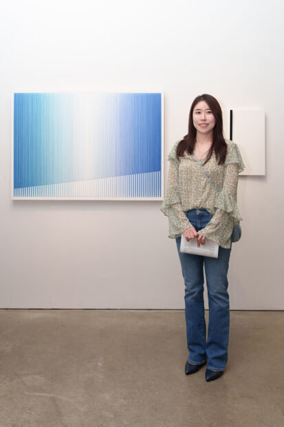 A photograph of artist Bumin Kim standing with her minimalist painting in a gallery.