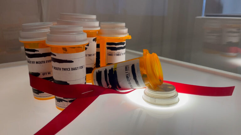 Detail image of Tricia Earl's found object piece - a group of used prescription bottles loosely tied with a red ribbon.