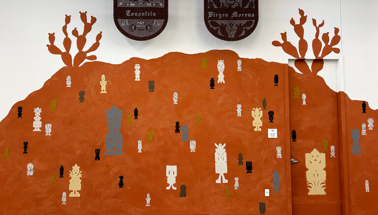 A detail photograph of a collaborative mixed media mural featuring a large red hill with cacti painted on a wall and cut-paper deities of varying sizes overlaid on the image. Two large, intricate cut-paper works hang above the hill.