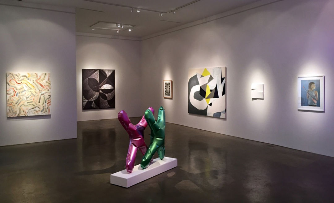 An art gallery show with paintings on the walls and two sculptures of X shapes made in colorful metal, resembling mylar balloons